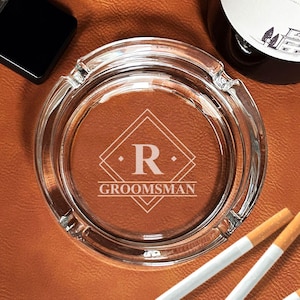 May include: A clear glass ashtray with a diamond-shaped design and the letter "R" in the center. The text "GROOMSMAN" is engraved below the letter. Three cigarettes are in the foreground.
