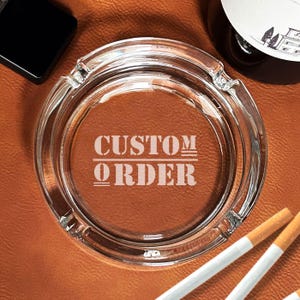 May include: A clear glass ashtray with the words "CUSTOM ORDER" etched on the bottom. The ashtray is sitting on a brown leather surface.