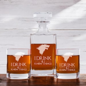 May include: A set of three clear glass whiskey glasses and a decanter with a white wolf graphic and the text "I drink & I know things".