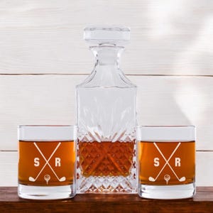 May include: Two clear glass whiskey tumblers with the initials "SR" and a golf club and ball design. A clear glass decanter with a diamond pattern is in the center.