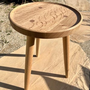 May include: A small, round wooden table with a natural wood finish. The table has three legs and a slightly raised edge around the top. The table is made of solid wood and has a rustic look.
