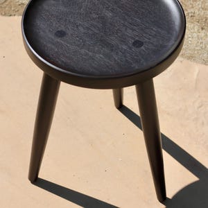 May include: A dark brown wooden stool featuring a circular seat with three visible circular joinery marks on the surface and three tapered legs extending downwards. The stool has a smooth, polished finish and a simple, functional design.