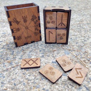 Peut inclure: Jeu de runes en bois avec une bo&icirc;te orn&eacute;e d'un motif de boussole et une autre bo&icirc;te contenant quatre runes. D'autres runes sont &eacute;parpill&eacute;es. Les runes et les bo&icirc;tes sont en bois brun clair.
