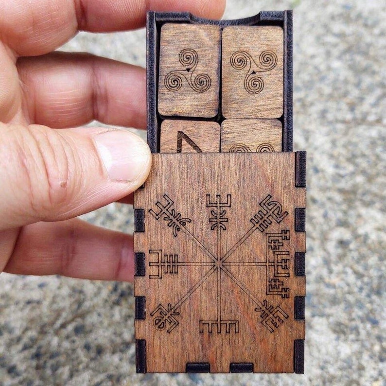 Peut inclure: Une bo&icirc;te en bois avec un couvercle coulissant, contenant des tuiles en bois avec des symboles grav&eacute;s. La bo&icirc;te pr&eacute;sente un motif en forme de boussole sur le devant. Les tuiles ont des motifs en spirale et g&eacute;om&eacute;triques. Le bois a une teinte brune chaude.