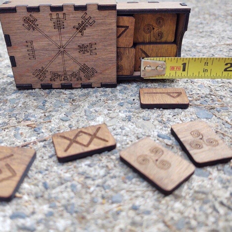 Peut inclure: Jeu de runes en bois avec une bo&icirc;te orn&eacute;e de symboles grav&eacute;s. La bo&icirc;te est ouverte, r&eacute;v&eacute;lant plusieurs runes. Certaines runes sont &eacute;parpill&eacute;es autour de la bo&icirc;te. Un m&egrave;tre ruban indique que la bo&icirc;te mesure environ 2,5 cm de large.