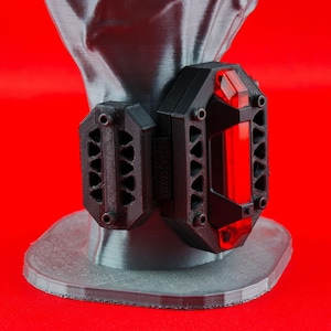 以下が含まれることがあります： 幾何学模様の黒と赤の3Dプリントの腕輪。腕輪は前面に赤い半透明のパネルが付いている。