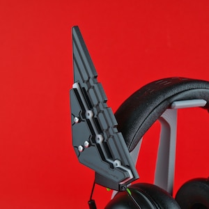 Puede incluir: Un accesorio para auriculares impreso en 3D de color negro con un diseño geométrico. El accesorio está diseñado para ajustarse sobre las almohadillas de los auriculares y tiene una superficie texturizada.