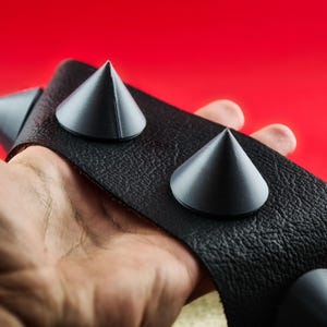 May include: Black leather wristband with large, gray conical studs. The wristband has a textured surface and is held against a red background. The studs are evenly spaced along the band.