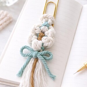 May include: A handmade macrame bookmark featuring a gold-colored paper clip. The bookmark has a knotted design in white, light blue, and brown, with a matching tassel and bow. The bookmark is resting on an open notebook.