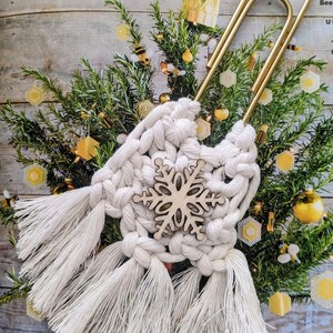 Puede incluir: Decoración de macramé blanco con un copo de nieve de madera en el centro, sujeta a un clip dorado. El macramé tiene flecos largos. El fondo incluye un pequeño árbol de hoja perenne con adornos amarillos y dorados.