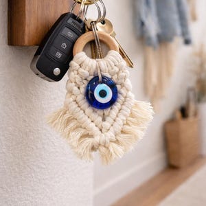 May include: A key holder with keys and a macrame keychain. The keychain features a wooden ring, cream-colored macrame, and a blue glass evil eye charm. The keys are attached to a gold-colored ring, along with a car key fob.