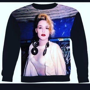 May include: A black sweatshirt with a printed image of a person with blonde hair wearing a white shirt and large black hoop earrings. The background is a blue and white patterned design.