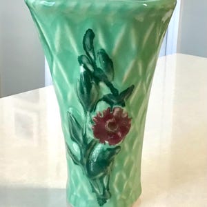 May include: A light green ceramic vase with a flared top and a textured diamond pattern across the surface. The front features a hand-painted design of a dark red flower with green leaves and stems, centered on the body of the vessel.