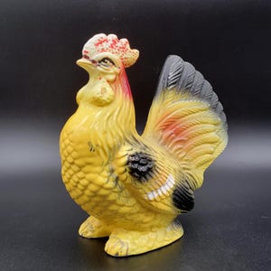 May include: A ceramic rooster figurine featuring a bright yellow body with textured feather details. The comb is painted with red speckles, while the tail feathers are a mix of black, yellow, and orange. The figure stands on a base.