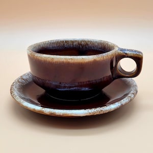 May include: A brown ceramic teacup and saucer set. The cup has a rounded shape with a large handle and sits on a matching saucer. The set has a glossy finish with a lighter brown rim.