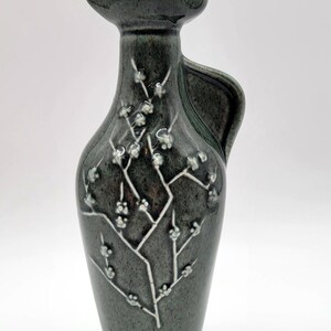 May include: A dark green ceramic decorative bottle with a matching stopper. The bottle features a white floral design and a handle. The bottle has a wide base and a narrow neck. The overall shape is reminiscent of a pitcher.