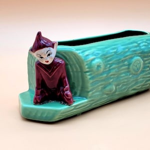 May include: A vintage ceramic planter shaped like a turquoise log, featuring a seated elf figurine. The elf wears a burgundy outfit and a pointed hat. The planter is rectangular with a hollow top for plants. A charming decorative piece.