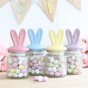 May include: Four glass jars with pastel coloured bunny ear lids filled with pastel coloured candy eggs. The jars are on a light wood surface.