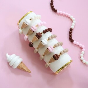 May include: A selection of ice cream cone bracelets and a necklace against a pink backdrop. The bracelets showcase different ice cream flavours, such as white, pink, and brown, with gold detailing. A single ice cream cone figurine is also visible.