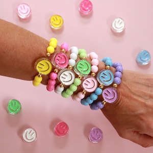 May include: A selection of colourful beaded bracelets with smiley face charms. The bracelets feature yellow, pink, white, green, blue, and purple beads. The charms are clear with a smiling face design.