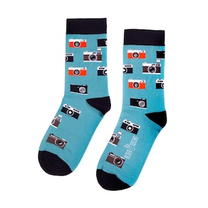 Op de afbeelding: Een paar novelty sokken met een teal achtergrond en een patroon van verschillende camera's in zwart, bruin en oranje. De sokken hebben zwarte hielen en tenen, en zwarte banden aan de bovenkant. De tekst "ALTER SOCKS" staat op de zijkant.