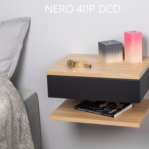 May include: A black floating bedside table with a drawer and a shelf. The bedside table has a light wood top and is mounted to a white wall. There are two decorative blocks on the top shelf, one grey and one pink.
