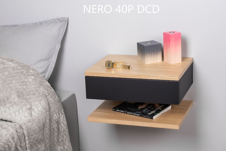 May include: A black floating bedside table with a drawer and a shelf. The bedside table has a light wood top and is mounted to a white wall. There are two decorative blocks on the top shelf, one grey and one pink.