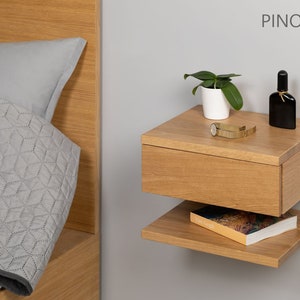 May include: A light wood floating nightstand with a drawer and a shelf. The nightstand is mounted to a wall and has a small plant, a watch, and a bottle of perfume on top. The text "PINO 30 P D" is visible on the wall.