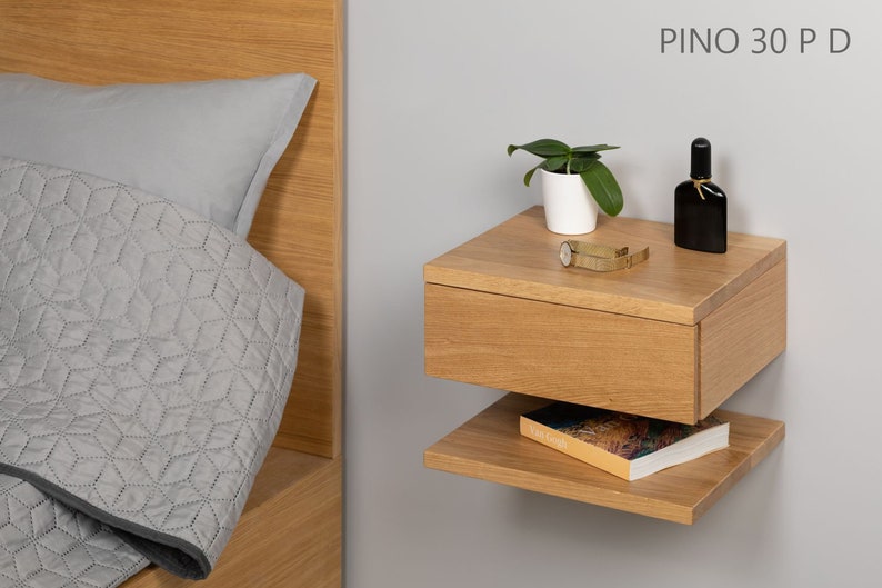 May include: A light wood floating nightstand with a drawer and a shelf. The nightstand is mounted to a wall and has a small plant, a watch, and a bottle of perfume on top. The text "PINO 30 P D" is visible on the wall.
