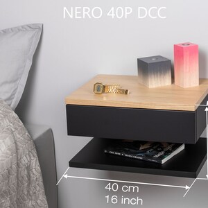 May include: A black floating bedside table with a drawer and a lower shelf. The nightstand is 40 cm wide, 23 cm deep, and 30 cm tall. It has a wood top and is shown with a watch, two decorative boxes, and a book on the shelf.