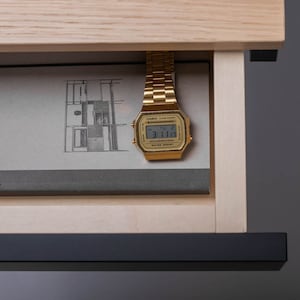 May include: A gold-tone digital watch with a black face and a black band is placed in a wooden drawer with a book. The watch face displays the time 3:11.