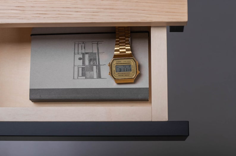May include: A gold-tone digital watch with a black face and a black band is placed in a wooden drawer with a book. The watch face displays the time 3:11.