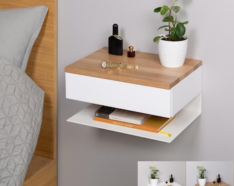 May include: White floating bedside table with a drawer and a shelf. The bedside table has a wood top and is mounted to the wall. A small plant, perfume bottles, and a watch are on the top of the bedside table.