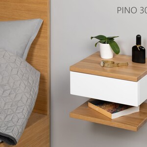 May include: A white floating nightstand with a drawer and a lower shelf. The nightstand is made of wood with a light wood finish. The nightstand is mounted to a wall and has a small plant, a watch, and a bottle of perfume on top.
