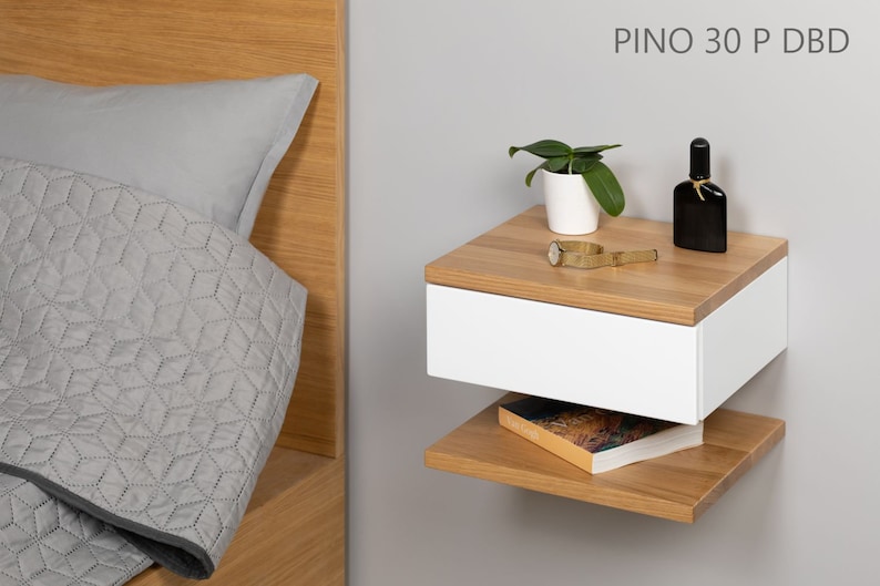 May include: A white floating nightstand with a drawer and a lower shelf. The nightstand is made of wood with a light wood finish. The nightstand is mounted to a wall and has a small plant, a watch, and a bottle of perfume on top.