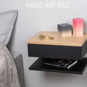 May include: A black floating bedside table with a drawer and a shelf. The bedside table has a wood top and is mounted to a wall. There are two decorative blocks on the top of the bedside table, one is grey and the other is pink. A gold watch is on the top of the bedside table. The text "NERO 40P DCC" is visible on the wall above the bedside table.