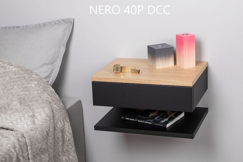 May include: A black floating bedside table with a drawer and a shelf. The bedside table has a wood top and is mounted to a wall. There are two decorative blocks on the top of the bedside table, one is grey and the other is pink. A gold watch is on the top of the bedside table. The text "NERO 40P DCC" is visible on the wall above the bedside table.