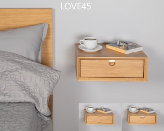May include: A light wood floating bedside table with a drawer and a small cup of coffee and a book on top. The bedside table is mounted to a white wall. LOVE45 is written in white text on the wall.