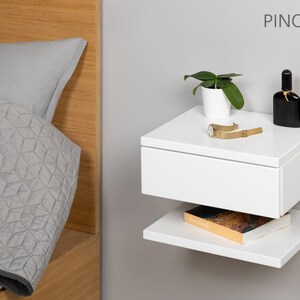 May include: White floating nightstand with a drawer and a lower shelf. The nightstand has a plant, a watch, and a bottle of perfume on top. The text "PINO 30 P B" is visible in the upper right corner of the image.