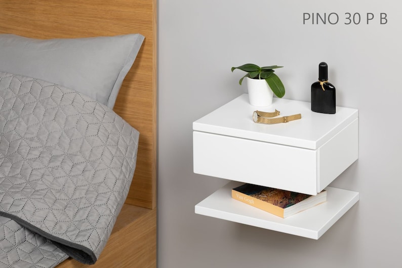 May include: White floating nightstand with a drawer and a lower shelf. The nightstand has a plant, a watch, and a bottle of perfume on top. The text "PINO 30 P B" is visible in the upper right corner of the image.