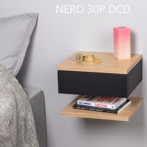 May include: A black floating bedside table with a drawer and a shelf. The bedside table has a light wood top and is mounted to a white wall. A pink candle, a watch, and a book are on the bedside table. The text "NERO 30P DCD" is printed on the wall above the bedside table.
