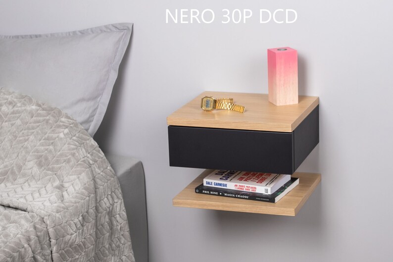 May include: A black floating bedside table with a drawer and a shelf. The bedside table has a light wood top and is mounted to a white wall. A pink candle, a watch, and a book are on the bedside table. The text "NERO 30P DCD" is printed on the wall above the bedside table.