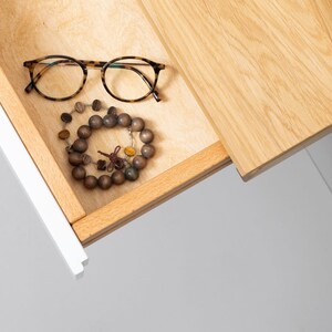 May include: A wooden drawer with a light wood finish is open, revealing a pair of tortoiseshell eyeglasses and a brown wooden beaded bracelet.