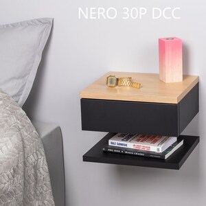 May include: A black floating bedside table with a drawer and a shelf. The bedside table has a wood top and is mounted to a white wall. A pink candle, a gold watch, and a book are on the bedside table. The text "NERO 30P DCC" is visible above the bedside table.