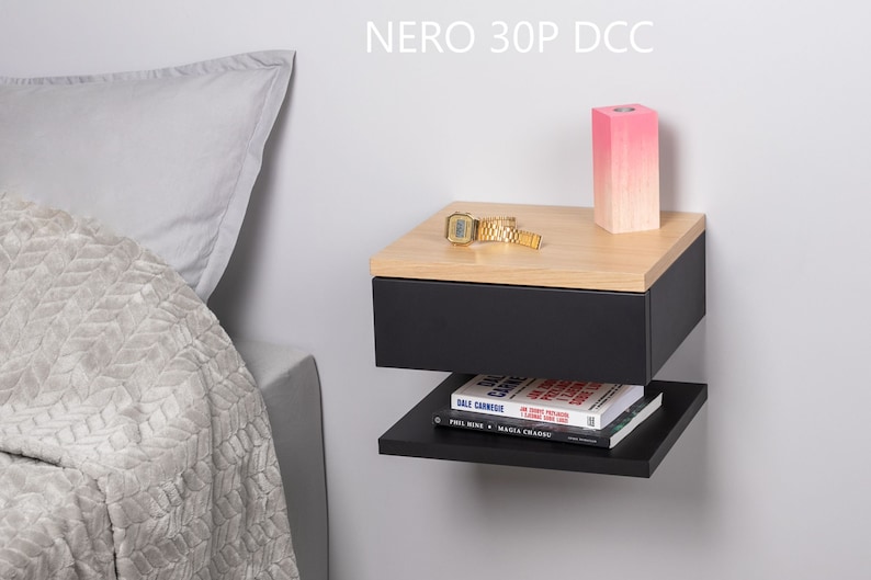 May include: A black floating bedside table with a drawer and a shelf. The bedside table has a wood top and is mounted to a white wall. A pink candle, a gold watch, and a book are on the bedside table. The text "NERO 30P DCC" is visible above the bedside table.