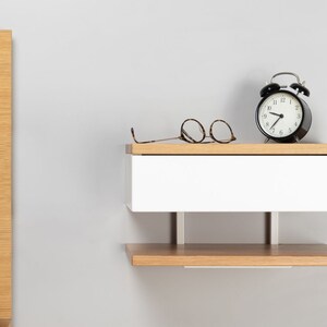 May include: A white floating nightstand with a wooden top and a lower shelf. The nightstand has a drawer and is mounted to the wall. A black alarm clock and a pair of glasses are on the top shelf.