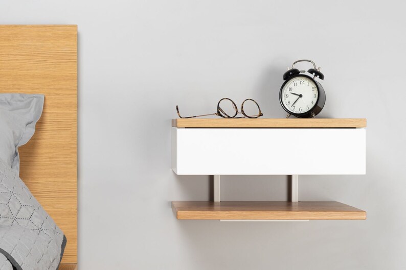 May include: A white floating nightstand with a wooden top and a lower shelf. The nightstand has a drawer and is mounted to the wall. A black alarm clock and a pair of glasses are on the top shelf.