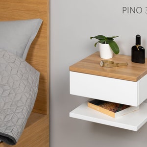 May include: White floating nightstand with a drawer and a shelf. The nightstand has a wood top and is mounted to a wall. A small plant, a watch, and a bottle of perfume are on the top of the nightstand. The text "PINO 30 P DB" is printed on the wall above the nightstand.