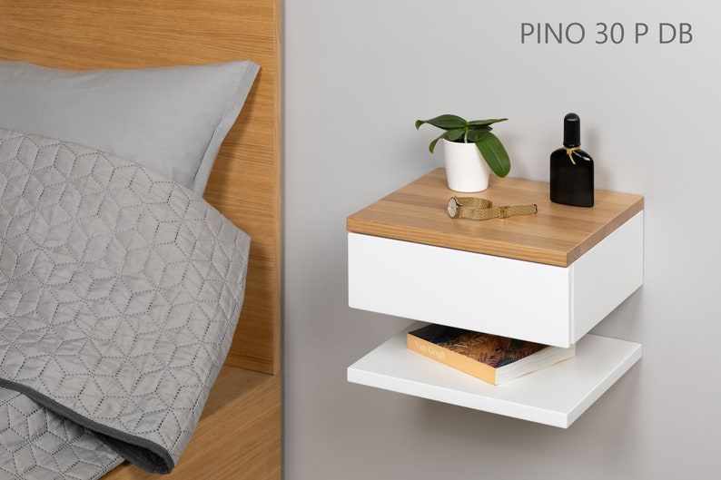 May include: White floating nightstand with a drawer and a shelf. The nightstand has a wood top and is mounted to a wall. A small plant, a watch, and a bottle of perfume are on the top of the nightstand. The text "PINO 30 P DB" is printed on the wall above the nightstand.
