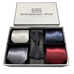 May include: A white gift box with a black lid containing a set of five ties and a bow tie. The ties are in the colors silver, gray, navy blue, and burgundy. The box has the text "MR MAHOGANY ROW - THE TUXEDO COLLECTION -" printed on the lid.
